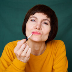 Obraz premium Smiling mature brunette woman painting lips with lipstick, close-up face portrait of middle-aged lady in bright yellow sweater on bright colorful dark green background. Generation X, 40s