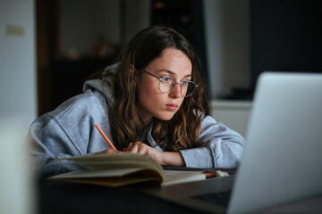 Young millennial woman at home look at laptop and make notes in notebook. Learn new skill from online classes or masterclass. New generation use technology for education and freelance