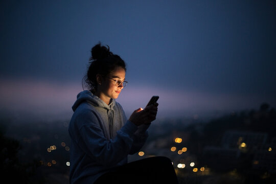 Beautiful young woman use smartphone at night. Millennial generation z female scroll through news feed on app on her phone. Cinematic film look photo of pretty woman at sunset use phone
