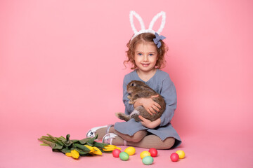 cute child girl in bunny ears holding rabbit, tulips, colorful eggs on pink background