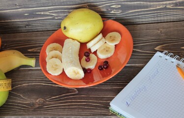 Cut the banana into slices on a platter, a notebook and fruit on a wooden table.
