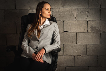 portrait of a beautiful, stylish, businesslike, confident businesswoman, brunette in a suit sitting on a leather chair and looking away, on a gray background. Low key. Selective focus