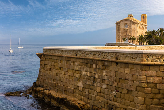 The sea wall and small church on the tiny island of Tabarca off the coast of Alicante in Spain. Two sailing boats are in the water and anchored. There are several palm trees on the right.