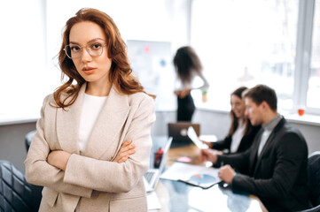 Creative modern office. Confident attractive redhead young woman, programmer or designer, stands in the office looks and smiles friendly at the camera, in the background colleagues are out of focus