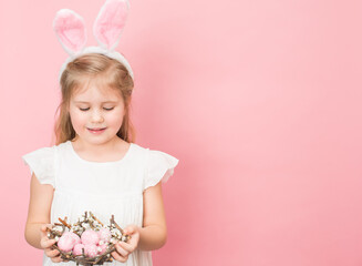Obraz na płótnie Canvas Happy little girl wearing bunny ears holding nest made of sticks full of painted eggs on pink background. Easter day children and fun concept.