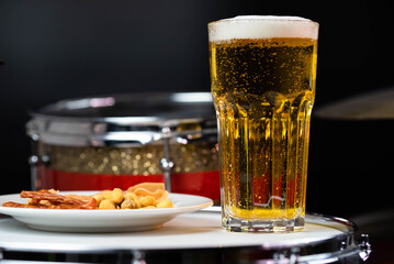 Glass of light beer with salty snacks on professional drum set closeup. Drumsticks, drums and cymbals, at live music rock concert, in the club stage, bar, or in recording studio