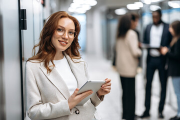 Creative modern office. Confident attractive redhead young woman, programmer or designer, stands in the office looks and smiles friendly at the camera, in the background colleagues are out of focus