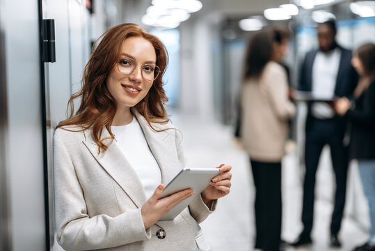 Creative Modern Office. Confident Attractive Redhead Young Woman, Programmer Or Designer, Stands In The Office Looks And Smiles Friendly At The Camera, In The Background Colleagues Are Out Of Focus