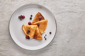 Top view, thin pancakes with berries on a plate on a gray textured background. Russian traditional pancakes. East Slavic traditional holiday Maslenitsa. Place to copy text.