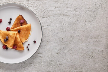 Top view, thin pancakes with berries on a plate on a gray textured background. Russian traditional pancakes. East Slavic traditional holiday Maslenitsa. Place to copy text.