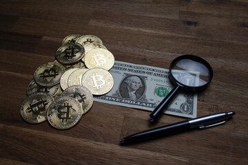 Pile of american dollars cash on brown wooden table. Next to it are several gold bitcoin digital cryptocurrency coins, a pen and a magnifying glass. Bank image and photo background.