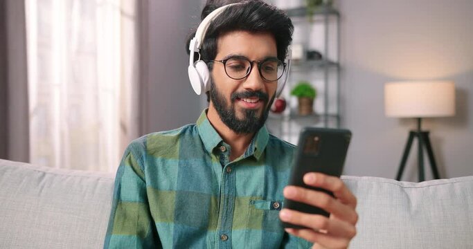 Close up portrait of joyful handsome young Indian guy listening to music in white headphones and surfing internet on smartphone watching videos in social network app sitting on sofa at home