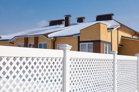 White Plastic Fence In A Modern Cottage Village On A Clear Winter Day. Snow Drifts In Front Of The Vinyl Fence Against A Blue Sky, The Roof Of The Cottage Is Covered With Snow