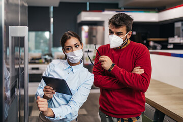 Young man talking with saleswoman about refrigerator he wants to buy. They are both with face protective masks. Pandemia, Covid-19 concept.