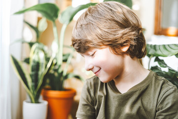 Portrait of Smiling child Kid boy in green t shirt with blue eyes in living room at sunny day. Stylish interior with a lot of plants. Home gardening, love of houseplants. Spring time. 