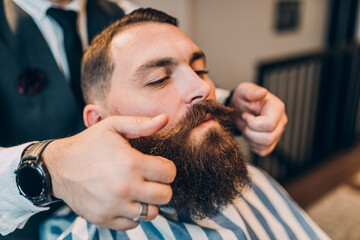 Young good looking hipster man visiting barber shop. Trendy and stylish beard styling and cut.