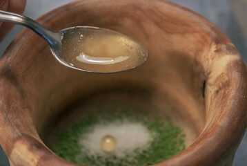 Close horizontal image of clay bowl full of matcha tea in powder with sugar while putting honey with a spoon on gray stone background. Matcha tea preparation 2021.