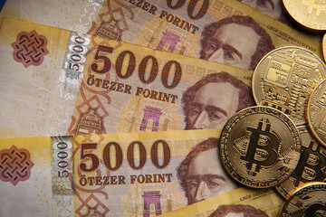 5000 Hungarian forint paper cash line. Next to it are a number of gold bitcoin (BTC) digital cryptocurrency coins. On a blue background.
