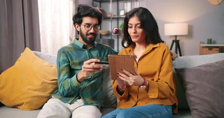 Portrait of happy cheerful young Indian couple boyfriend and girlfriend sitting on couch in cozy room at home buying stuff online paying with credit card on internet using tablet, shopping concept