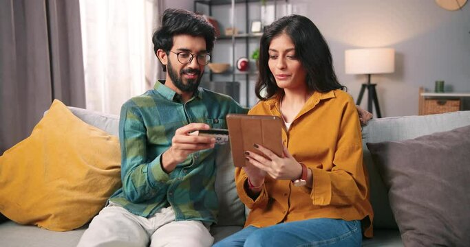 Portrait Of Happy Cheerful Young Indian Couple Boyfriend And Girlfriend Sitting On Couch In Cozy Room At Home Buying Stuff Online Paying With Credit Card On Internet Using Tablet, Shopping Concept