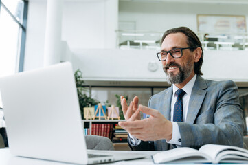 Fototapeta premium Satisfied mature businessman is having a video call with colleagues, discussing project. Handsome male employee uses laptop, communicate with coworkers, smiling
