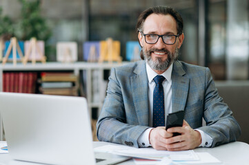 Portrait of a confident senior business man in formal suit and eyeglasses is sitting at the workplace, looking and friendly smiling directly at the camera - obrazy, fototapety, plakaty