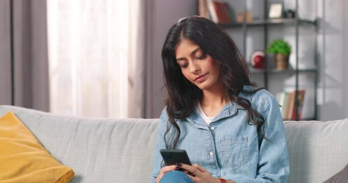 Portrait of Hindu happy beautiful young woman texting on smartphone resting on sofa in cozy room, female browsing online on cellphone in good mood and smiling, social network user concept