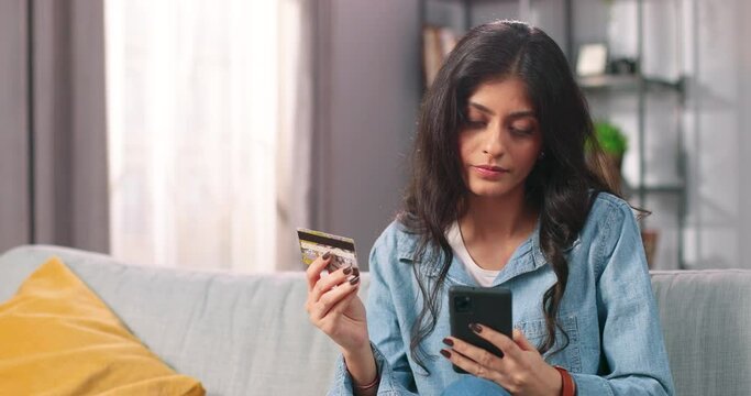 Close up portrait of happy beautiful Hindu young woman buying stuff online on sales on smartphone paying with credit card while sitting on sofa in cozy room at home, internet shopping concept