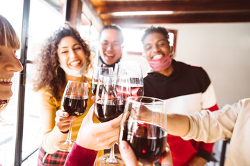 Happy friends wearing protective face masks toasting red wine at restaurant - New normal friendship concept with young people having fun at home party - Focus on glasses