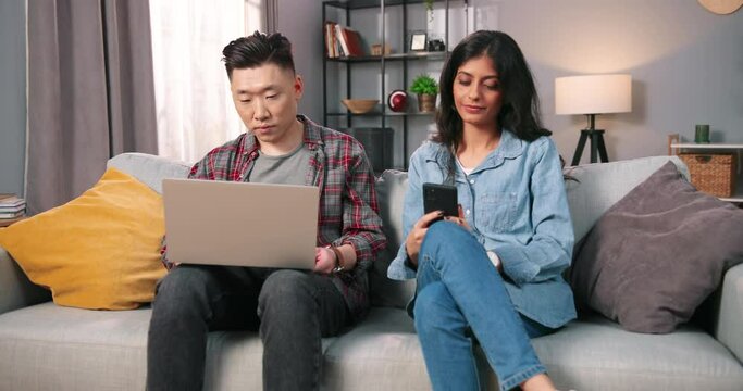 Multi-ethnic married couple sitting on couch in cozy living room at home using tech digital gadgets. Husband browsing on laptop while his wife searching internet, texting on smartphone leisure concept