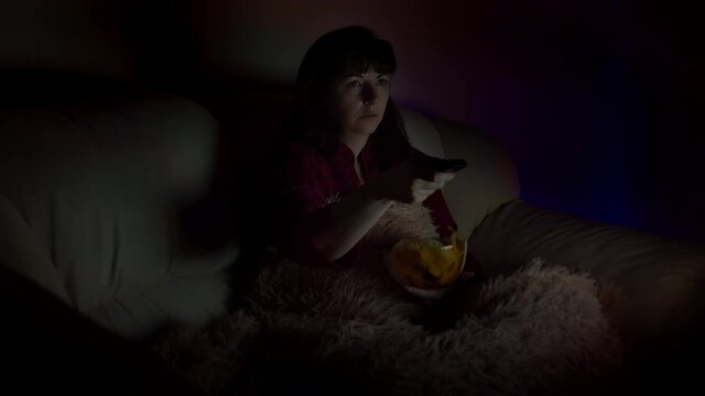 sad brunette woman sits in sofa with chips at night and watches tv with emotion