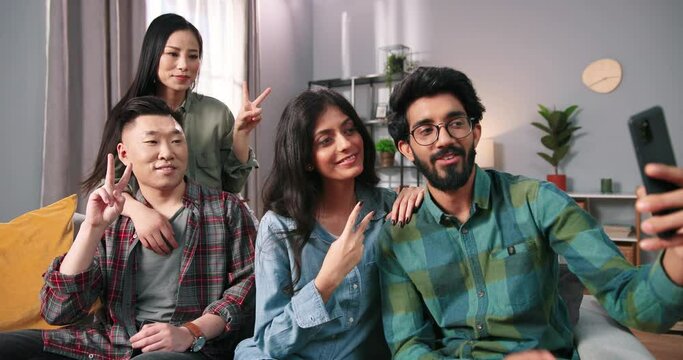 Portrait of mixed-race happy positive young Asian and Hindu couples sitting on sofa at home and posing smiling to smartphone camera taking selfie photos together, cellphone pictures, leisure concept