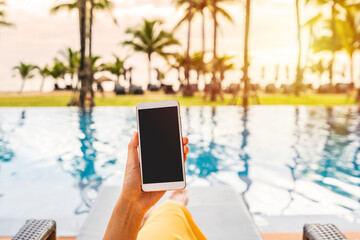 Young woman traveler relaxing and using a mobile phone by a hotel pool while traveling for summer...