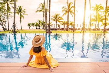 Young woman traveler relaxing and enjoying the sunset by a tropical resort pool while traveling for summer vacation - obrazy, fototapety, plakaty