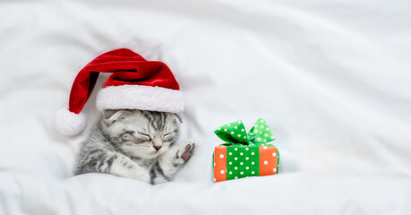 Cute kitten wearing red santa's hat sleeps with gift box under a white blanket on a bed. Top down view. Empty space for text
