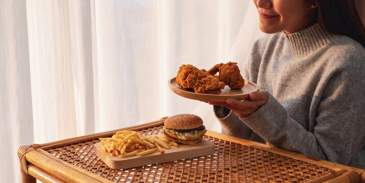 Closeup image of a young asian woman holding and eating fried chicken with hamburger and french fries on the table at home