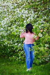 Naklejka na ściany i meble Portrait of a fat plump young woman with black hair and a blooming apple tree on a white floral background in a park on a spring day. Happy girl plus size.