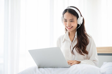 Happy beautiful young asian woman working on computer laptop and sitting on bed in house. She making video conference webinar and wearing headphone