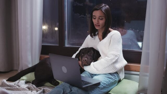 Young woman freelancer is sitting on the floor and petting the dog, working on a laptop, types text on the keyboard, evening time, remote work from country house, winter time.