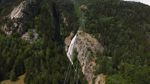 Electric Cables Over The Alpine Mountain With Green Forest And Waterfall On The Background In Italy. aerial
