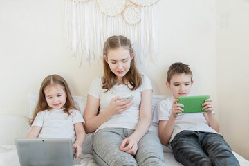 Three children of different ages are sitting on a bed with different electronic gadgets. A little girl sits with a laptop, a teenage girl with a phone, a boy with a glider.