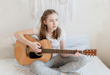 A teenage girl sits on a bed and plays an acoustic guitar.