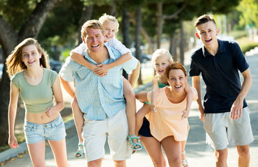 Obraz premium Positive man and woman with four kids running and carrying kids on their back in town