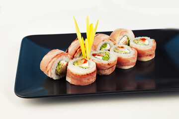 Sushi roll with bacon and fried sea bass on a black plate, ingredients fried sea bass, cream cheese, bacon, Iceberg salad, flying fish roe, Teriyaki sauce, rice, nori. For the restaurant menu.