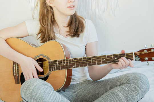 A young girl in a white T-shirt sits and plays an acoustic guitar.