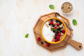 Breakfast, unsweetened yogurt with baked granola, raspberries and blueberries in a yellow bowl on a wooden board on a light concrete background. Breakfast recipes.