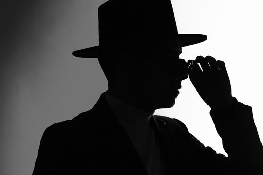 Silhouette of a man in a jacket and classial hat fixing sunglasses over white and grey background. Side view.