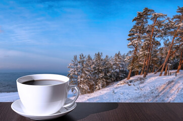 Obraz na płótnie Canvas Cup of coffee on the table with view of snowy beach, Baltic sea and covered in snow fir and pine trees on the hill.
