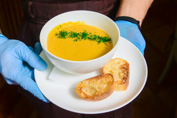 Squash pumpkin cream soup with fresh greenery served in a white bowl by waiter in restaurant or diner.
