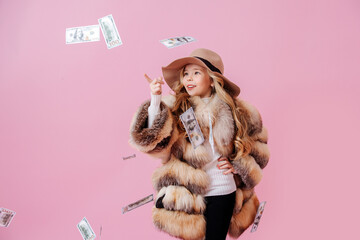 Pretty preteen girl in oversized luxurious fur coat and pole hat pointing finger at money raining at her. Over dark pink background.